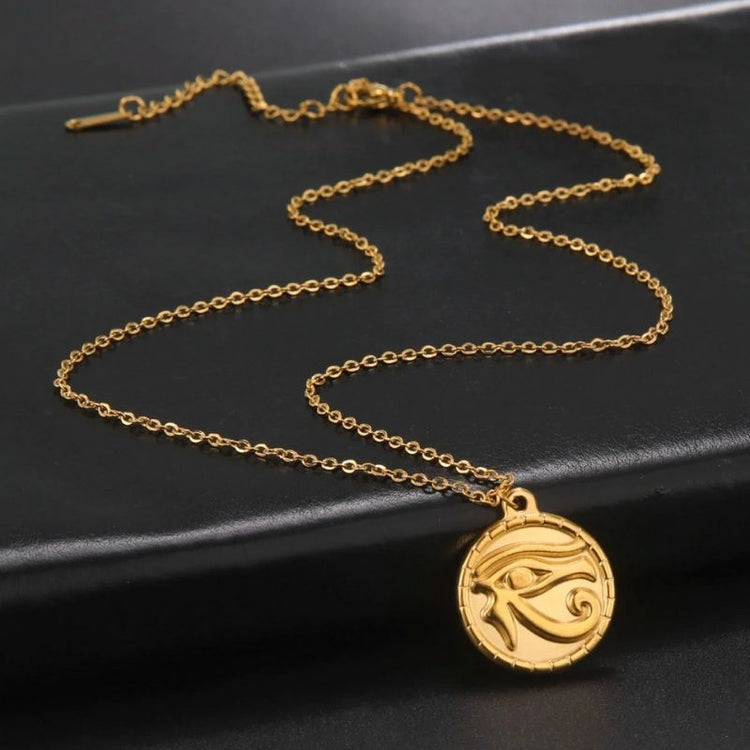 Pendentif Œil d'Horus en acier inoxydable couleur or, présenté avec sa chaîne délicate sur une surface noire, associant design mystique et élégance raffinée.