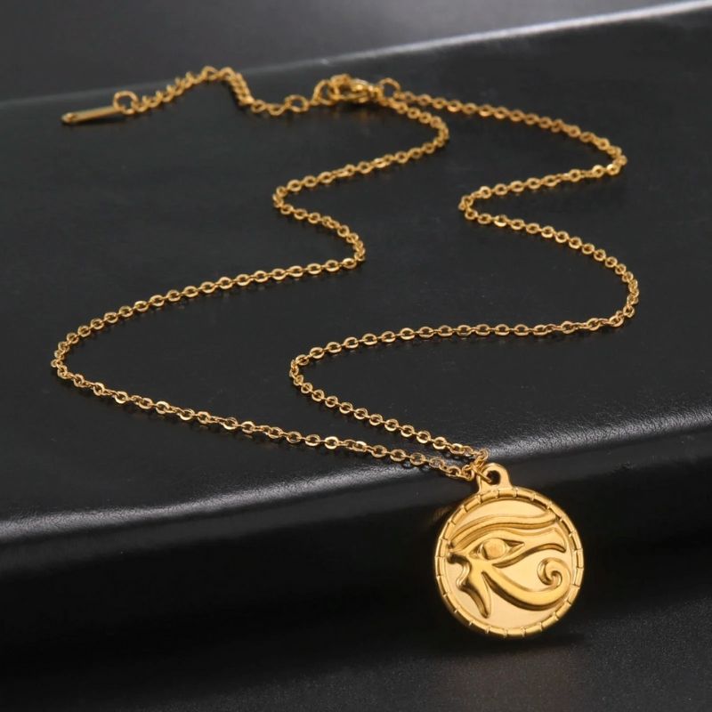 Pendentif Œil d'Horus en acier inoxydable couleur or, présenté avec sa chaîne délicate sur une surface noire, associant design mystique et élégance raffinée.