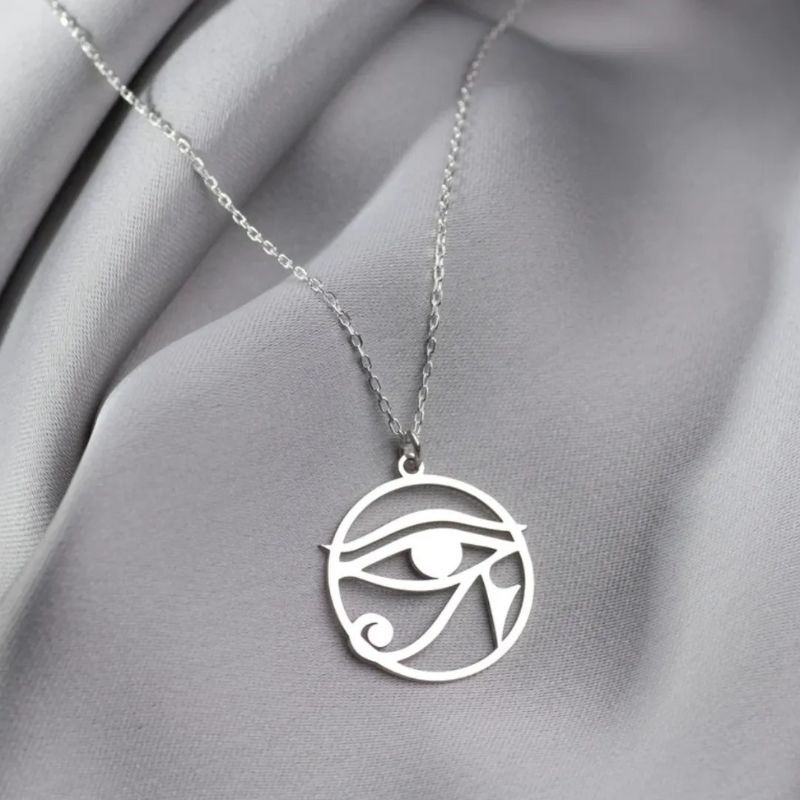 Collier avec pendentif Oeil d'Horus en argent, accroché à une chaîne fine, exposé sur un tissu gris soyeux.