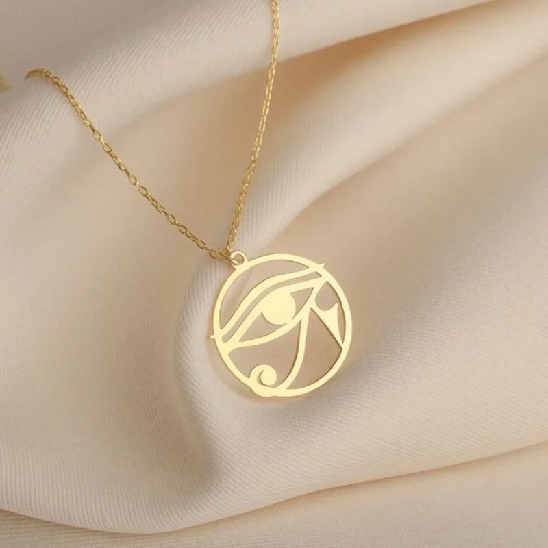 Pendentif Oeil d'Horus en or suspendu à une chaîne fine, présenté sur un tissu beige.