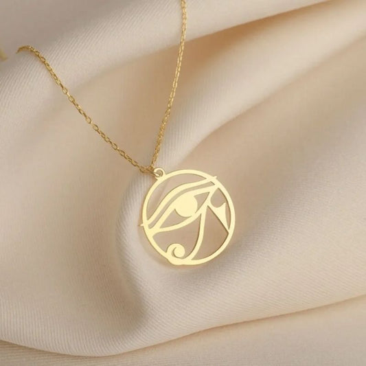Pendentif Oeil d'Horus en or suspendu à une chaîne fine, présenté sur un tissu beige.
