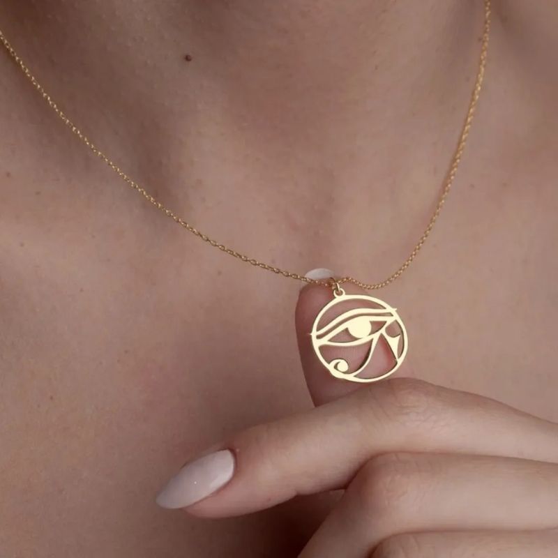 Pendentif Oeil d'Horus en or délicatement tenu entre les doigts d'une femme, illustrant son élégance et sa symbolique spirituelle.