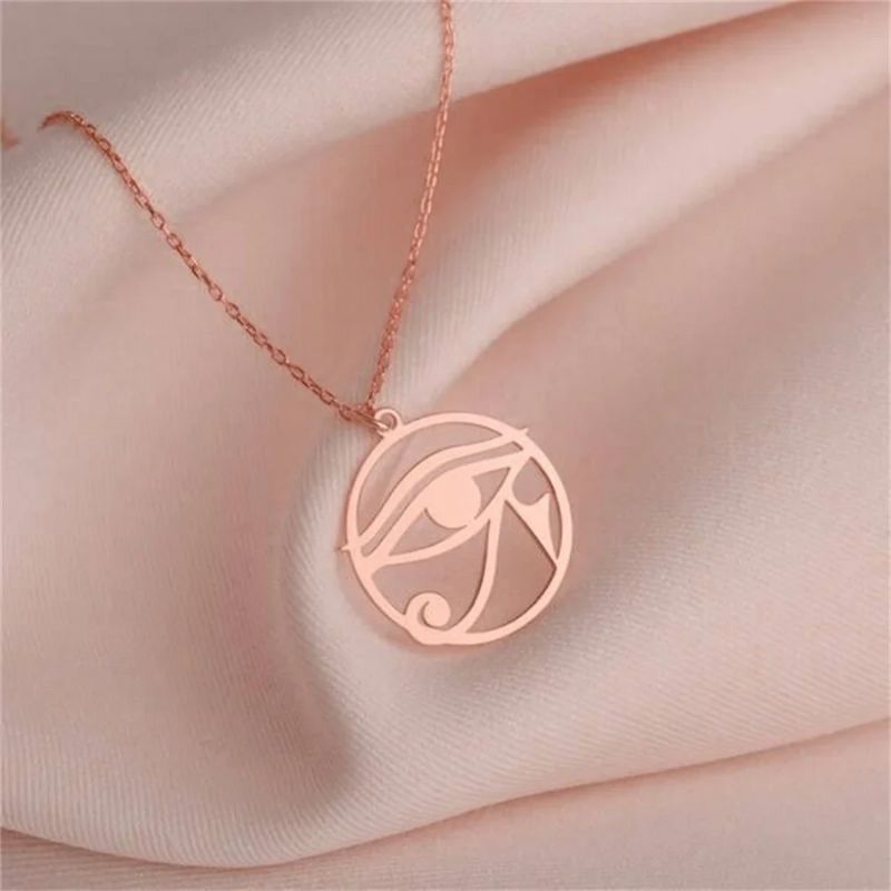 Pendentif Oeil d'Horus en or rose sur une chaîne délicate, posé sur un tissu rose poudré.