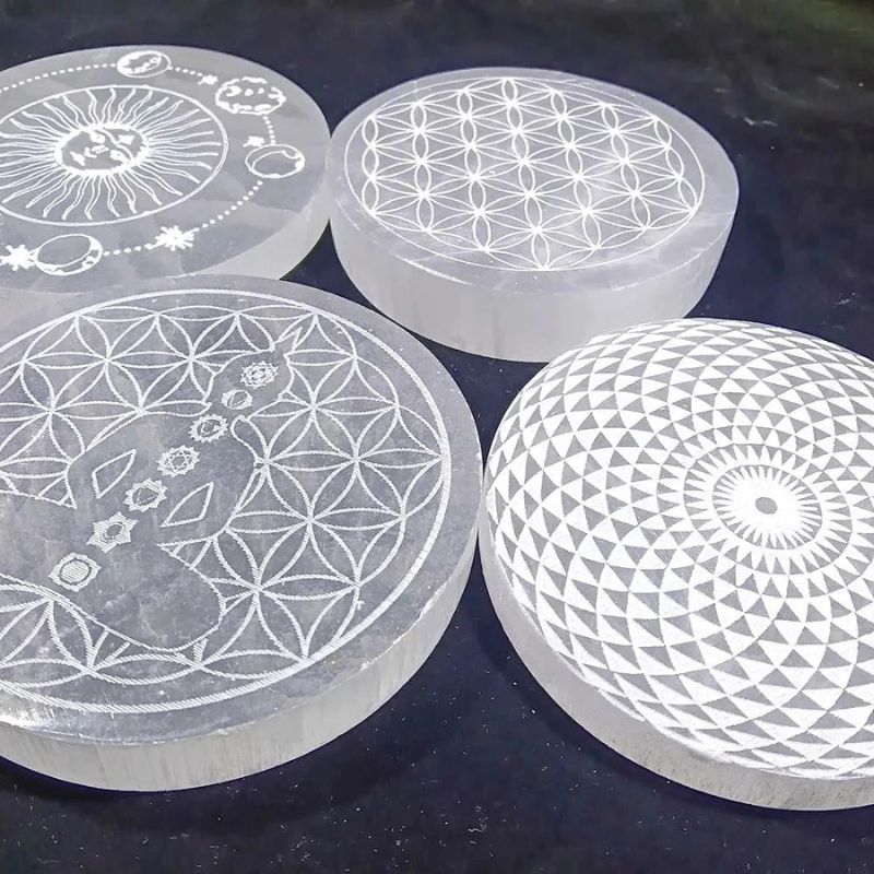 Plaques de rechargement en Sélénite Fleur de vie de 10cm avec différents motifs gravés, disposées sur fond noir.