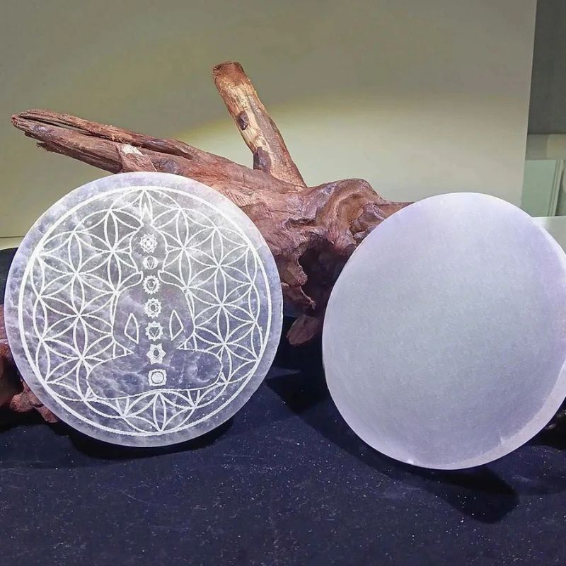 Plaque de rechargement Sélénite Fleur de vie et plaque lisse, disposées sur fond noir avec élément décoratif en bois.